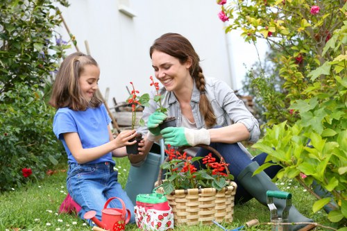 A woman and a young girl are engaged in gardening in a lush outdoor space in Thamesmead, with vibrant green plants and flowering bushes surrounding them. The woman, wearing a light grey jacket and green gardening gloves, is smiling as she holds a small potted plant with red flowers, while the girl, dressed in a blue t-shirt and denim shorts, carefully handles the plant. They are seated on a well-maintained grass area, with gardening tools, including a small watering can and a patterned basket, placed beside them. The garden features a variety of bushes and flowering plants, creating a colourful and natural environment. The weather appears bright and clear, supporting outdoor gardening activities, which are consistent with professional gardening services offered in Thamesmead. The scene highlights the enjoyment of gardening and outdoor maintenance, typical of residential garden landscapes in the Thamesmead area.