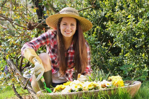 A young woman with long brown hair, smiling and wearing a straw hat, gardening gloves, and a red checkered shirt, is kneeling in a lush garden. She is tending to blooming yellow and white flowers in a large, circular wooden planter, surrounded by green foliage, shrubs, and small trees. The garden surface is composed of well-maintained grass, and in the background, there are densely packed leaves and branches from various plants. The weather appears sunny with natural light illuminating the scene, creating a bright and inviting outdoor environment that exemplifies typical gardening activities in Thamesmead. This setting highlights gardening and outdoor maintenance services offered by Gardening Services Thamesmead, focusing on floral care and garden beautification within a vibrant, leafy landscape.
