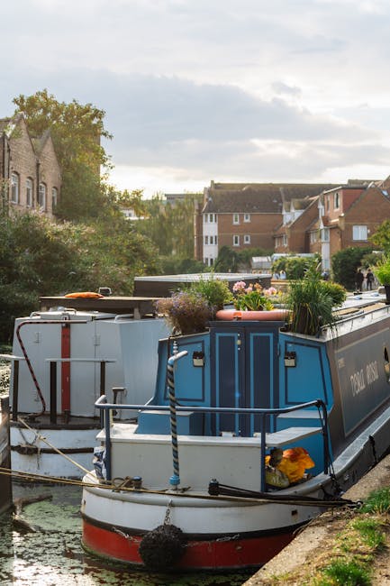 A tranquil scene along Thamesmead's canalside gardens featuring two narrowboats moored under tall, leafy willow trees that cascade over the water. The canal’s surface reflects the boats and surroundin