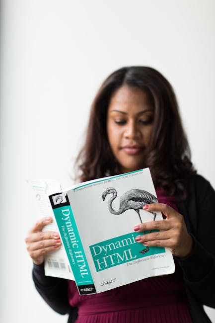 The image shows a woman with dark hair wearing a black blazer and a maroon top, holding an open book titled 'Dynamic HTML' with a cover illustration of a flamingo and technical text. She appears to be