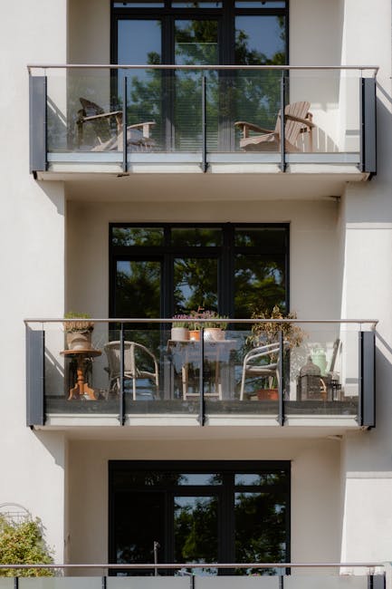 A three-storey balcony garden in a modern residential building, showcasing glass-panelled railings with black metal supports. The lower balcony features potted plants, a small table surrounded by four